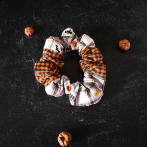 May include: A fabric hair scrunchie featuring a patchwork design of orange and black buffalo plaid fabric alternating with a white fabric printed with small Halloween motifs including pumpkins, bats, and moons, surrounded by three small orange pumpkins.