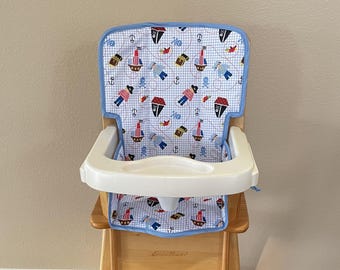 May include: A wooden high chair featuring a light-colored wood frame and a white plastic tray. The seat cushion is covered in a white fabric with a blue grid pattern, decorated with colorful pirate-themed illustrations including ships, anchors, and figures.