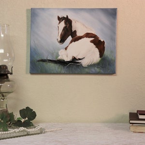 May include: A painting of a brown and white horse lying down in a field of green grass. The horse is looking to the left of the image. The painting is on a canvas and has a light blue background.