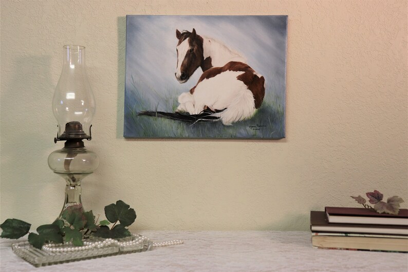 May include: A painting of a brown and white horse lying down in a field of green grass. The horse is looking to the left of the image. The painting is on a canvas and has a light blue background.
