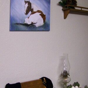 May include: A painting of a brown and white horse lying down on a blue background. The horse is looking to the left. The painting is framed and hanging on a white wall.