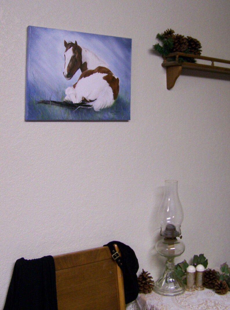 May include: A painting of a brown and white horse lying down on a blue background. The horse is looking to the left. The painting is framed and hanging on a white wall.