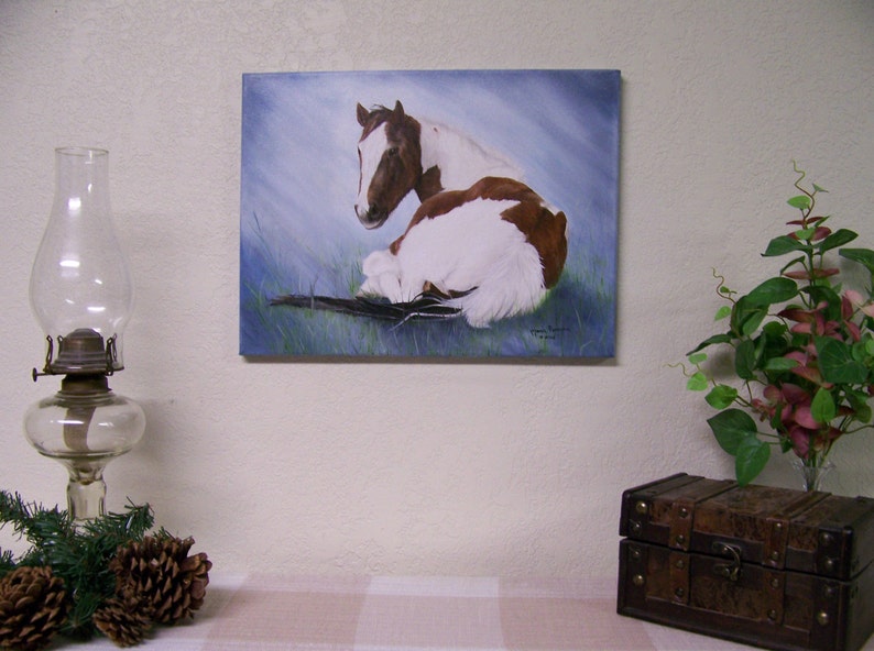 May include: A painting of two brown and white horses lying down in a field. The horses are facing away from the viewer, with the horse in the foreground resting its head on the back of the horse behind it. The painting has a blue sky background with a light blue and green field.