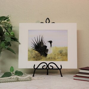 May include: A framed watercolor painting of a black and white bird with its tail feathers fanned out. The bird is standing in a grassy field. The painting is on a black easel.