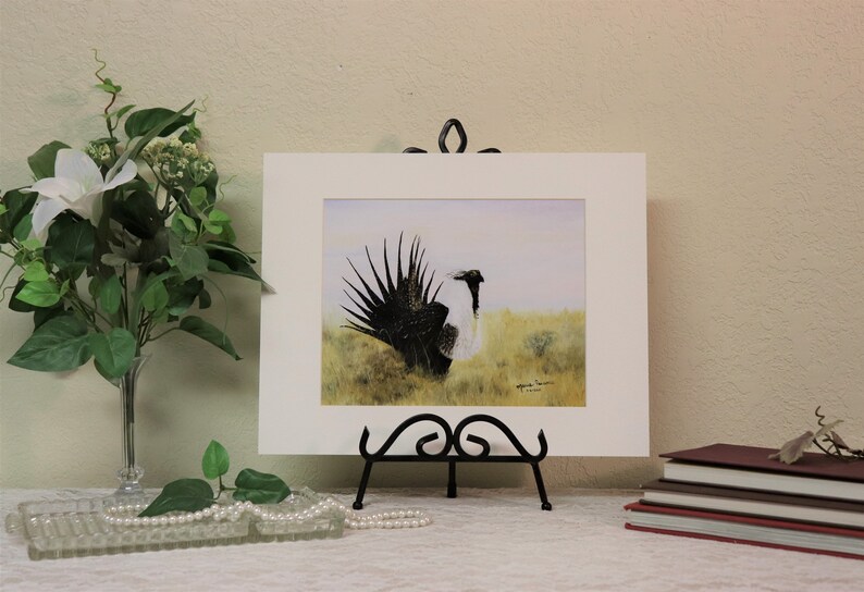May include: A framed watercolor painting of a black and white bird with its tail feathers fanned out. The bird is standing in a grassy field. The painting is on a black easel.