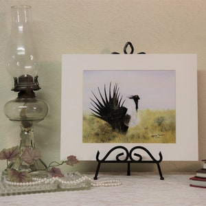 May include: A framed watercolor painting of a black and white bird with a long, feathery tail. The bird is standing in a field of green grass. The painting is signed by the artist, "Marie Irwin, 2018".