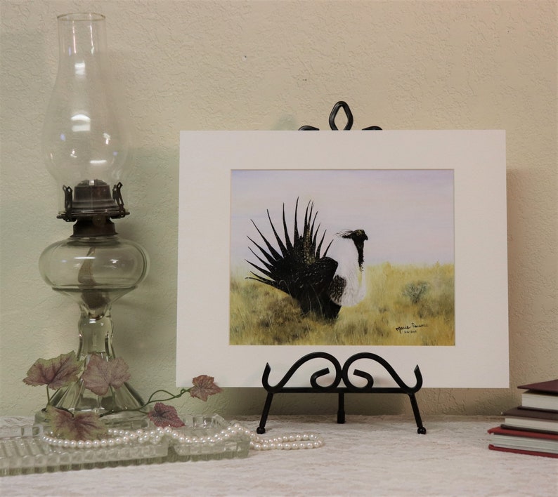 May include: A framed watercolor painting of a black and white bird with a long, feathery tail. The bird is standing in a field of green grass. The painting is signed by the artist, "Marie Irwin, 2018".
