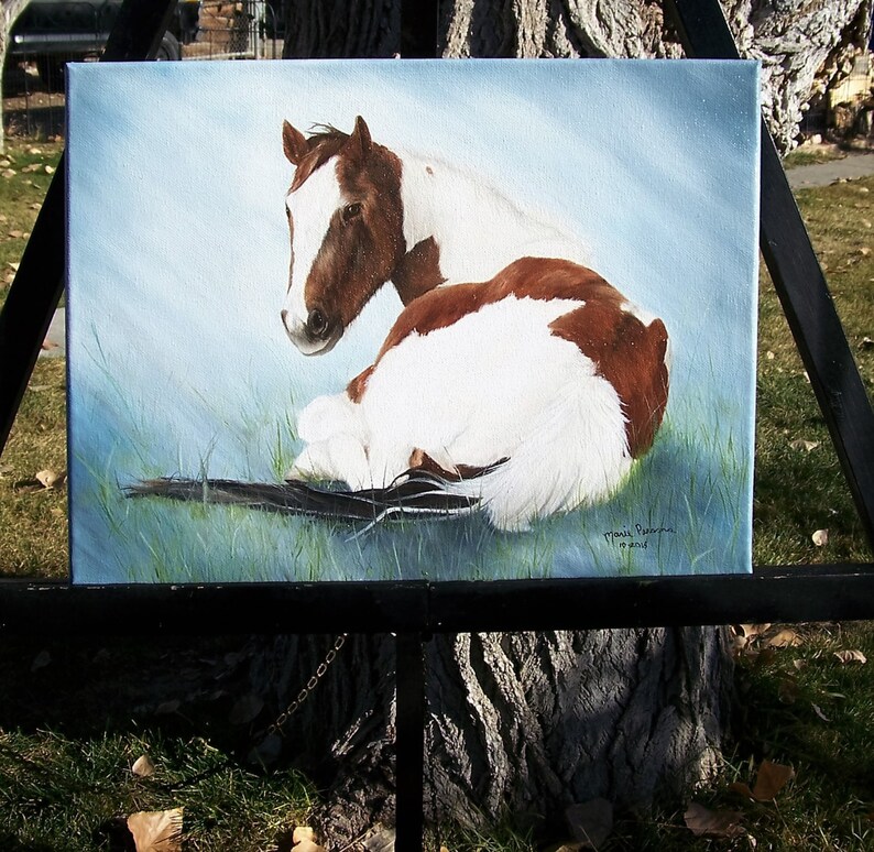 May include: A painting of a brown and white horse lying down in a field of green grass. The horse is looking to the left of the image. The painting is signed "Marie Robinson" in the bottom right corner.