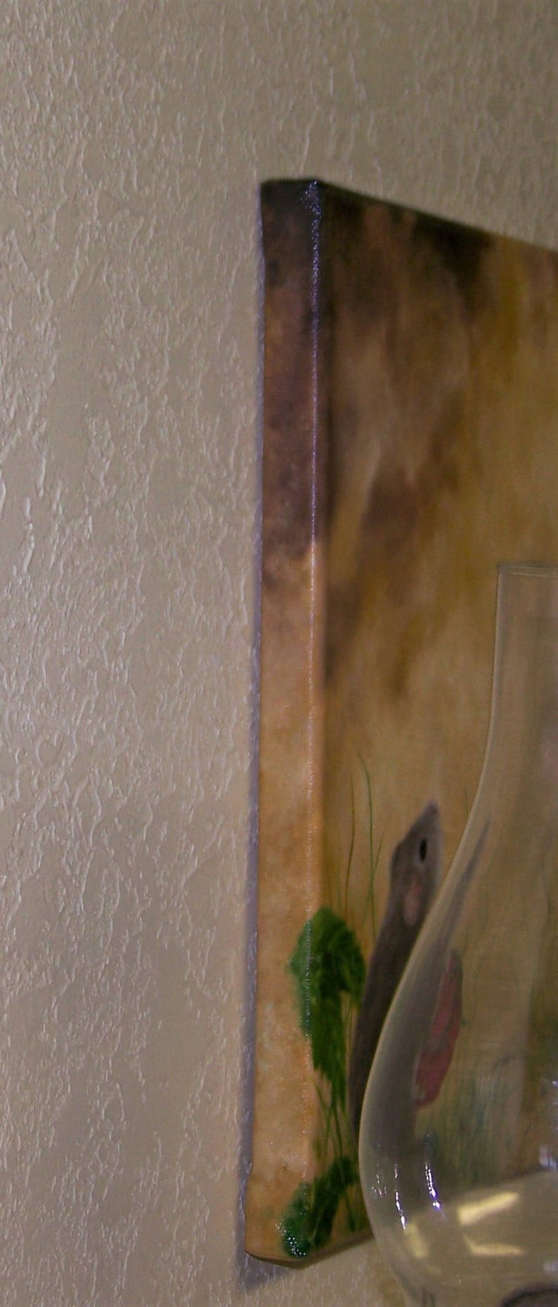Puede incluir: Un primer plano de una pintura sobre lienzo que presenta un peque&ntilde;o rat&oacute;n marr&oacute;n con plantas verdes y un jarr&oacute;n de vidrio transparente. La pintura tiene un fondo marr&oacute;n con un borde marr&oacute;n claro.