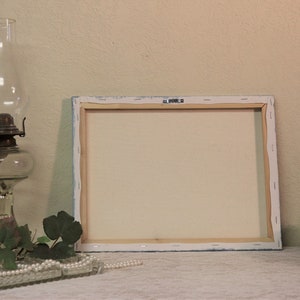May include: A blank canvas painting on a wooden frame, with a white background and a light blue border. The canvas is sitting on a white table with a glass oil lamp, green leaves, and a string of pearls.