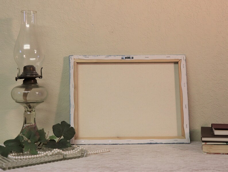 May include: A blank canvas painting on a wooden frame, with a white background and a light blue border. The canvas is sitting on a white table with a glass oil lamp, green leaves, and a string of pearls.