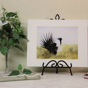 May include: A framed watercolor painting of a black and white bird with a long, feathery tail. The bird is standing in a field of green grass. The painting is signed by the artist.