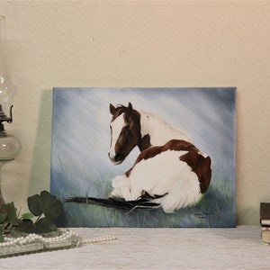 May include: A painting of a brown and white horse lying down in a field. The horse is looking to the left of the painting. The background is a light blue sky with white clouds.