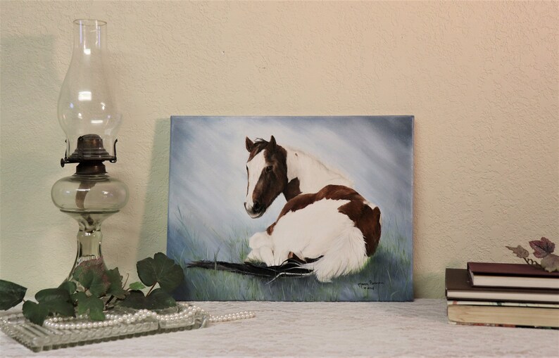 May include: A painting of a brown and white horse lying down in a field. The horse is looking to the left of the painting. The background is a light blue sky with white clouds.