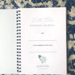 May include: A white spiral bound journal with the title "Little Blue Wedding Journal" and a space to write the names of the couple and the date of the wedding. The journal has a blue watercolour illustration of hydrangeas and the name "Winnefred Austin" with a website address.