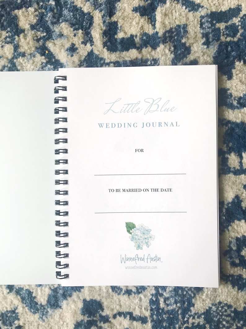 May include: A white spiral bound journal with the title "Little Blue Wedding Journal" and a space to write the names of the couple and the date of the wedding. The journal has a blue watercolour illustration of hydrangeas and the name "Winnefred Austin" with a website address.