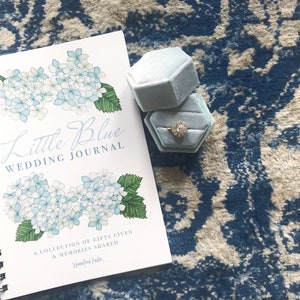 May include: A white spiral bound journal with a blue and white floral design and the text "Little Blue Wedding Journal" on the cover. The journal is on a blue and white patterned rug with a blue velvet ring box with a gold ring inside.