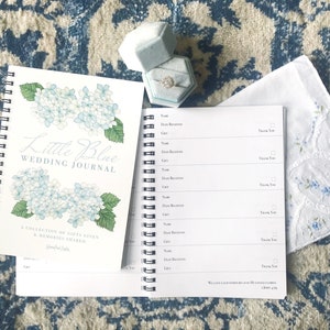May include: A white wedding journal with a blue hydrangea illustration on the cover. The journal is titled "Little Blue Wedding Journal" and has the text "A collection of gifts given & memories shared". A blue ring box with a diamond ring is on the right side of the image.