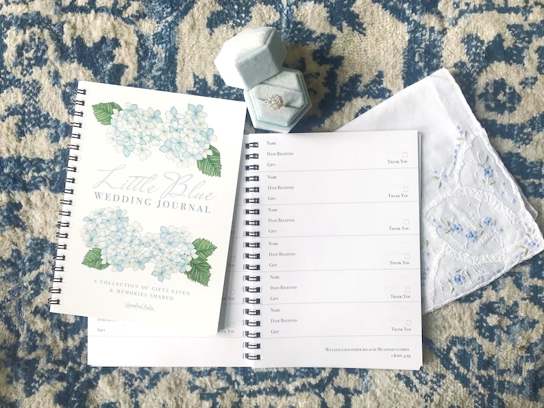 May include: A white wedding journal with a blue hydrangea illustration on the cover. The journal is titled "Little Blue Wedding Journal" and has the text "A collection of gifts given & memories shared". A blue ring box with a diamond ring is on the right side of the image.