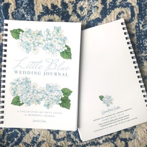 May include: A white spiral bound journal with a blue and white floral design on the cover. The title "Little Blue Wedding Journal" is written in cursive. The text "A collection of gifts given & memories shared" is printed below the title. The back cover features a smaller floral design and the text "Waenefred Austin SOMETHING BLUE Keep a record of wedding gifts, shower memories & planning details in your little blue journal."