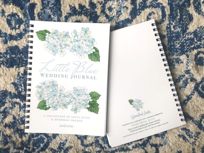May include: A white spiral bound journal with a blue and white floral design on the cover. The title "Little Blue Wedding Journal" is written in cursive. The text "A collection of gifts given & memories shared" is printed below the title. The back cover features a smaller floral design and the text "Waenefred Austin SOMETHING BLUE Keep a record of wedding gifts, shower memories & planning details in your little blue journal."