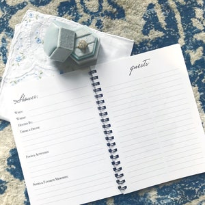 May include: A white notebook with a blue spiral binding. The notebook has lined pages and the words "Shower:" and "Guests" written on the front and back covers.