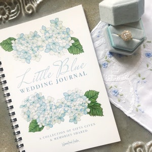 May include: A white spiral bound journal with a blue watercolour illustration of hydrangeas and the text "Little Blue Wedding Journal". The text below the illustration reads "A collection of gifts given & memories shared" and is signed "Winifred Austin."