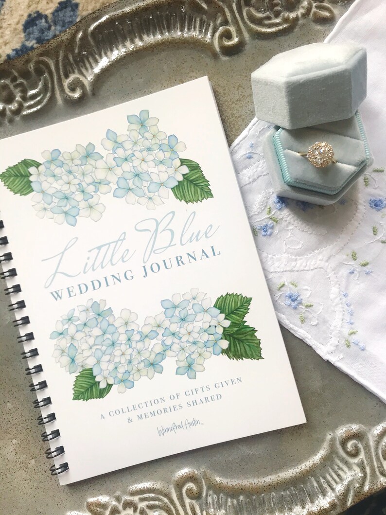 May include: A white spiral bound journal with a blue watercolour illustration of hydrangeas and the text "Little Blue Wedding Journal". The text below the illustration reads "A collection of gifts given & memories shared" and is signed "Winifred Austin."