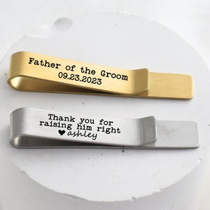 May include: Two engraved tie clips. The gold tie clip says "Father of the Groom 09.23.2023". The silver tie clip says "Thank you for raising him right ♥ ashley".