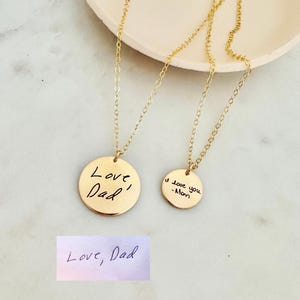 Könnte beinhalten: Zwei goldfarbene Scheibenhalsketten mit handschriftlichen Inschriften. Auf einer steht "Love, Dad" und auf der anderen "I love you - Mom". Eine kleine rechteckige Karte darunter sagt auch "Love, Dad". Die Halsketten liegen auf einer hellen Oberfläche.