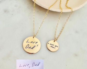 Puede incluir: Dos collares de disco dorados con inscripciones manuscritas. Uno dice "Love, Dad" y el otro "I love you - Mom". Una pequeña tarjeta rectangular debajo también dice "Love, Dad". Los collares están sobre una superficie clara.