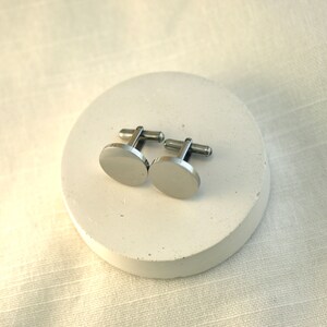 May include: A pair of silver cufflinks with a round, smooth design. The cufflinks are on a white circular surface.