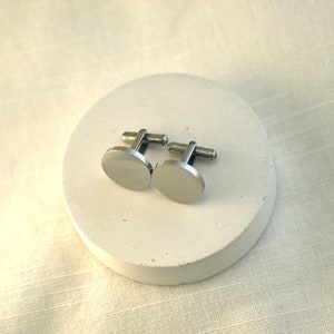 May include: A pair of silver cufflinks with a round, smooth design. The cufflinks are on a white circular surface.
