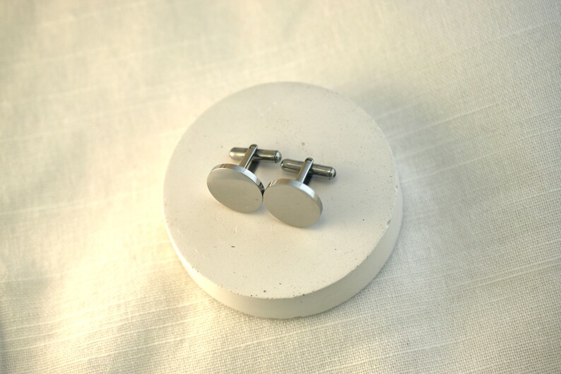 May include: A pair of silver cufflinks with a round, smooth design. The cufflinks are on a white circular surface.