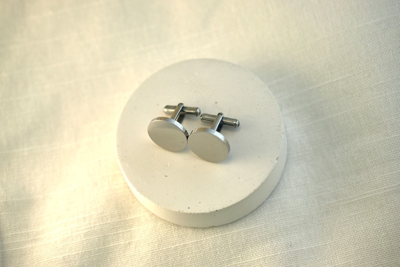 May include: A pair of silver cufflinks with a round, smooth design. The cufflinks are on a white circular surface.