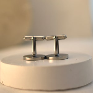 May include: A pair of silver cufflinks with a simple design. The cufflinks are on a white surface.