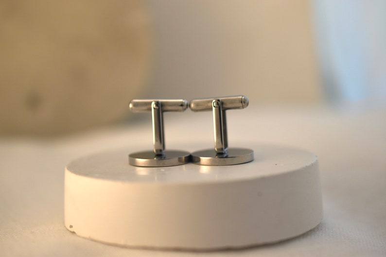 May include: A pair of silver cufflinks with a simple design. The cufflinks are on a white surface.