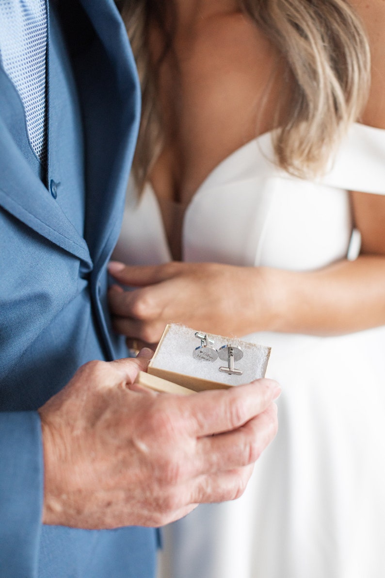 May include: A close-up of a small, rectangular box with two silver cufflinks inside. The cufflinks are round and have a design on them. The box is open and the cufflinks are visible.