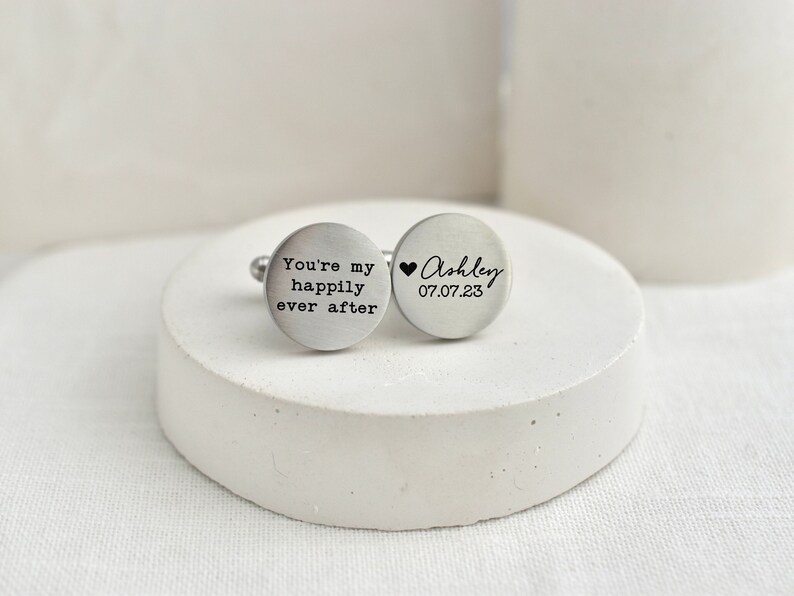 May include: Two silver cufflinks with engraved text. One cufflink says "You're my happily ever after". The other cufflink says "Ashley 07.07.23" with a small heart symbol.