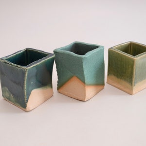 以下が含まれることがあります： 薄い茶色の土台と緑色の釉薬をかけた、小さな正方形のセラミックプランターが3つあります。釉薬は不均一で、独特の外観を生み出しています。