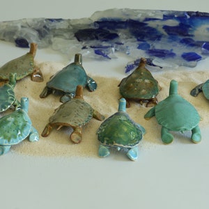 Peut inclure: Collection de figurines de tortues en céramique de différentes nuances de vert et de bleu, disposées sur une surface sablonneuse. Une pièce décorative en verre ressemblant à des vagues est en arrière-plan. Les tortues ont des détails et une finition brillante.