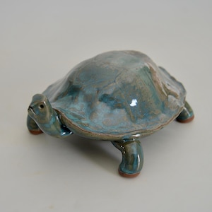 May include: A ceramic turtle figurine with a teal and brown glaze. The turtle has a detailed shell and is posed on four small legs. The figurine is a decorative item.