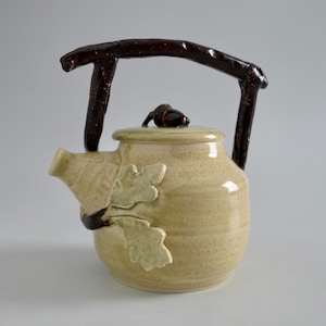 May include: A ceramic teapot with a twig handle and an acorn finial. The teapot is decorated with green oak leaves and has a light brown glaze.