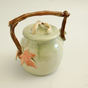 May include: A ceramic teapot with a twig handle and leaf accents. The teapot is glazed in a light green color and has a brown twig handle. The teapot has a leaf design on the side and a leaf-shaped lid.