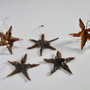 Puede incluir: Cinco adornos marrones en forma de estrella con una textura rústica similar a la madera. Cada adorno tiene una cuerda dorada para colgar.