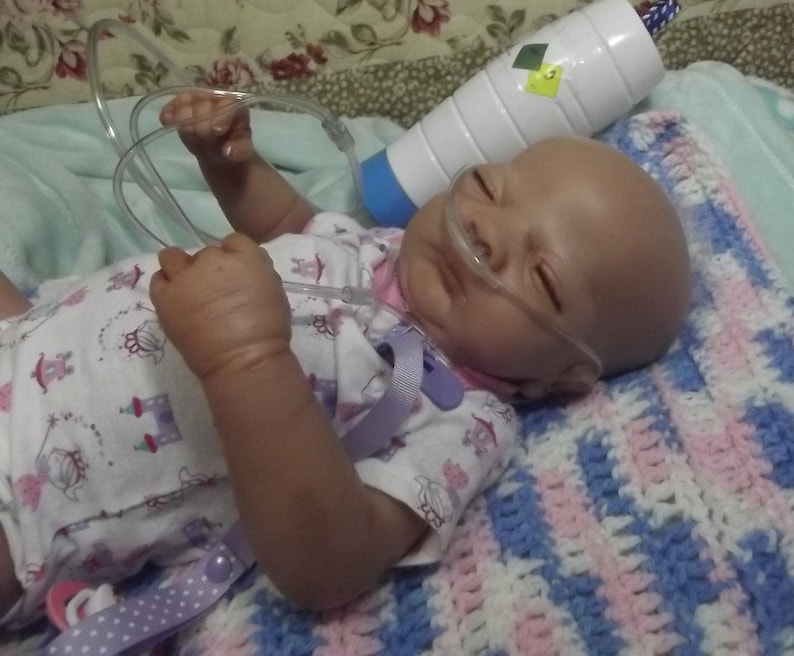 May include: A baby doll wearing a white and pink dress with a floral pattern, lying on a blue and pink striped blanket. The doll has a clear nasal cannula inserted in its nostrils and is connected to a clear tubing.