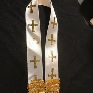 Może przedstawiać: Biała stola z satynowej wstążki z powtarzającym się wzorem metalicznych złotych krzyży nadrukowanych wzdłuż obu pionowych krawędzi. Dolne końce wstążki są wykończone ozdobną frędzlą w kolorze złotego metalu, kt&oacute;ra dodaje całości tekstury.