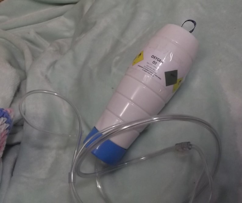 May include: A white oxygen tank with a blue cap and a clear plastic tube. The tank has a yellow warning label that says "Oxygen O2".