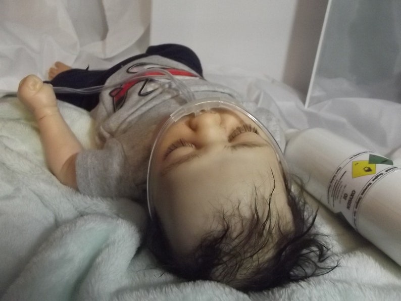 May include: A baby doll with dark hair and closed eyes is lying on a light blue blanket. The doll is wearing a gray shirt with a red design and a clear oxygen mask. A white oxygen tank with a yellow warning label is visible in the background.