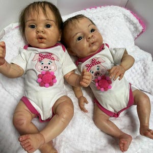 May include: Two realistic baby dolls are lying on a white blanket. Each doll wears a white onesie with pink trim and a pig graphic. The dolls have detailed facial features and are posed with their arms and legs slightly bent.